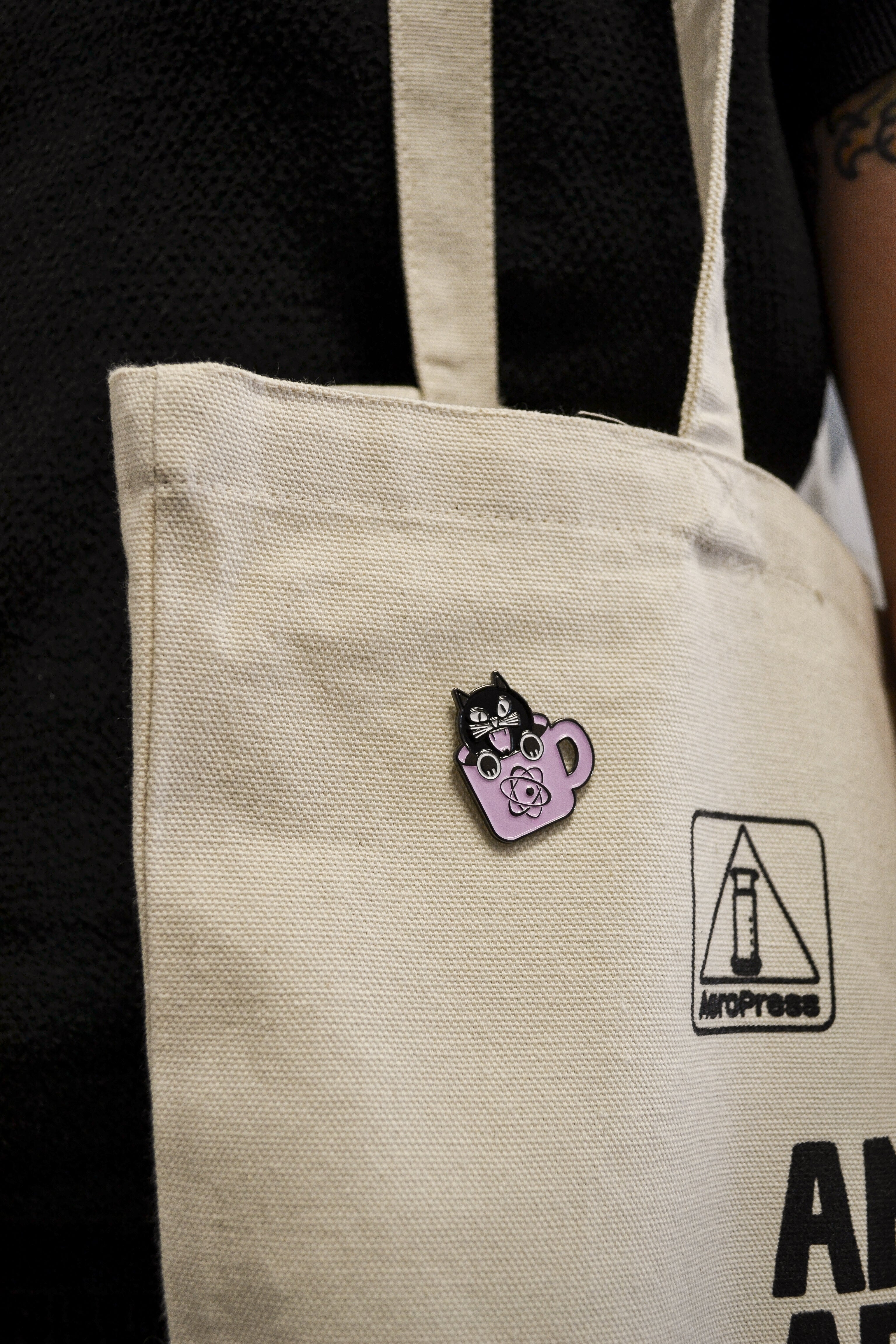 A close-up of a beige canvas tote bag worn over the shoulder, featuring a small enamel pin of a black cat in a purple teacup and a black square logo with the word 'MYPRESS'.