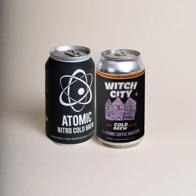 Two 12 oz cans, one black can that says "Atomic Nitro Cold Brew" and one black can that says "Witch City Cold Brew" with Halloween artwork.