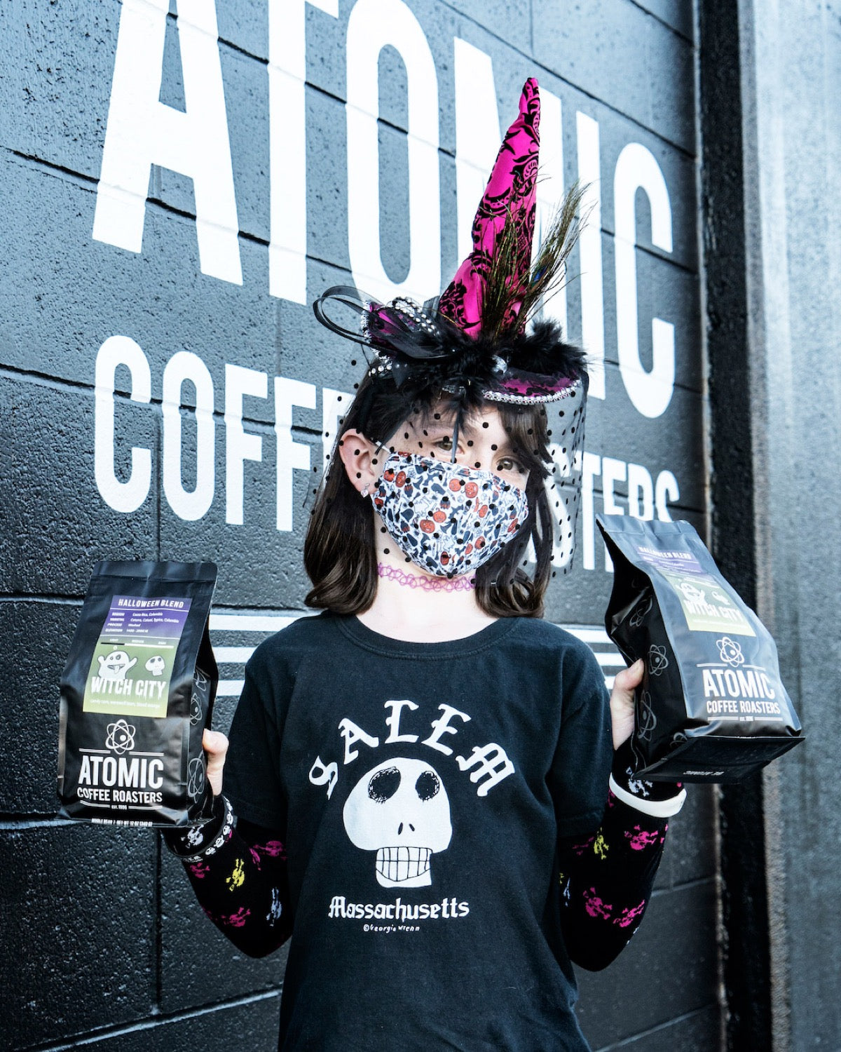 Georgia Wrenn in a witch hat outside the Atomic Coffee Roasters roastery, holding two bags of the first Witch City.
