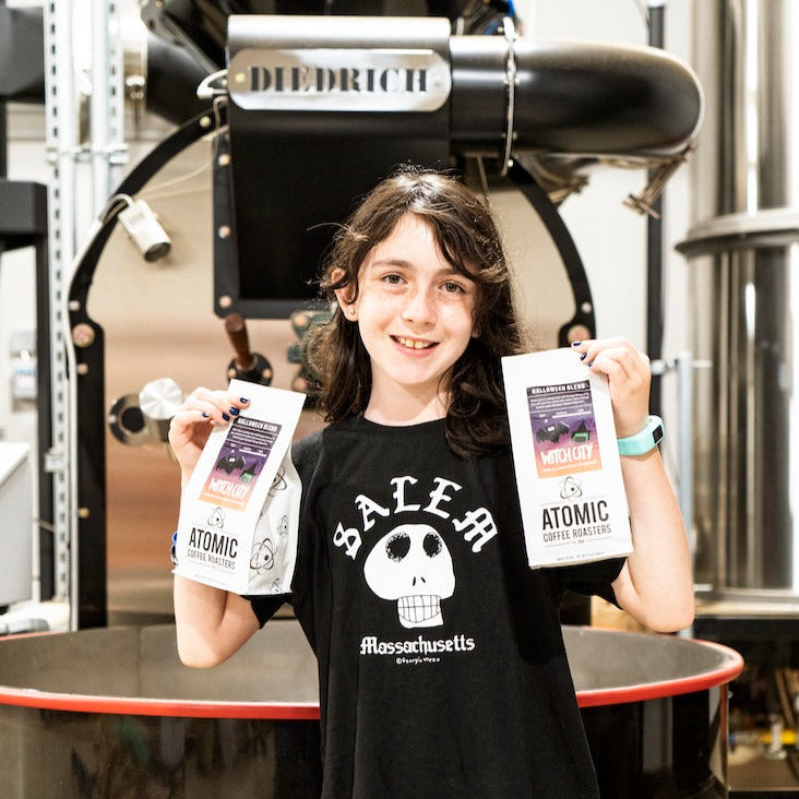 Georgia Wrenn at the roastery in front of the Diedrich roaster, holding two bags of Witch City.
