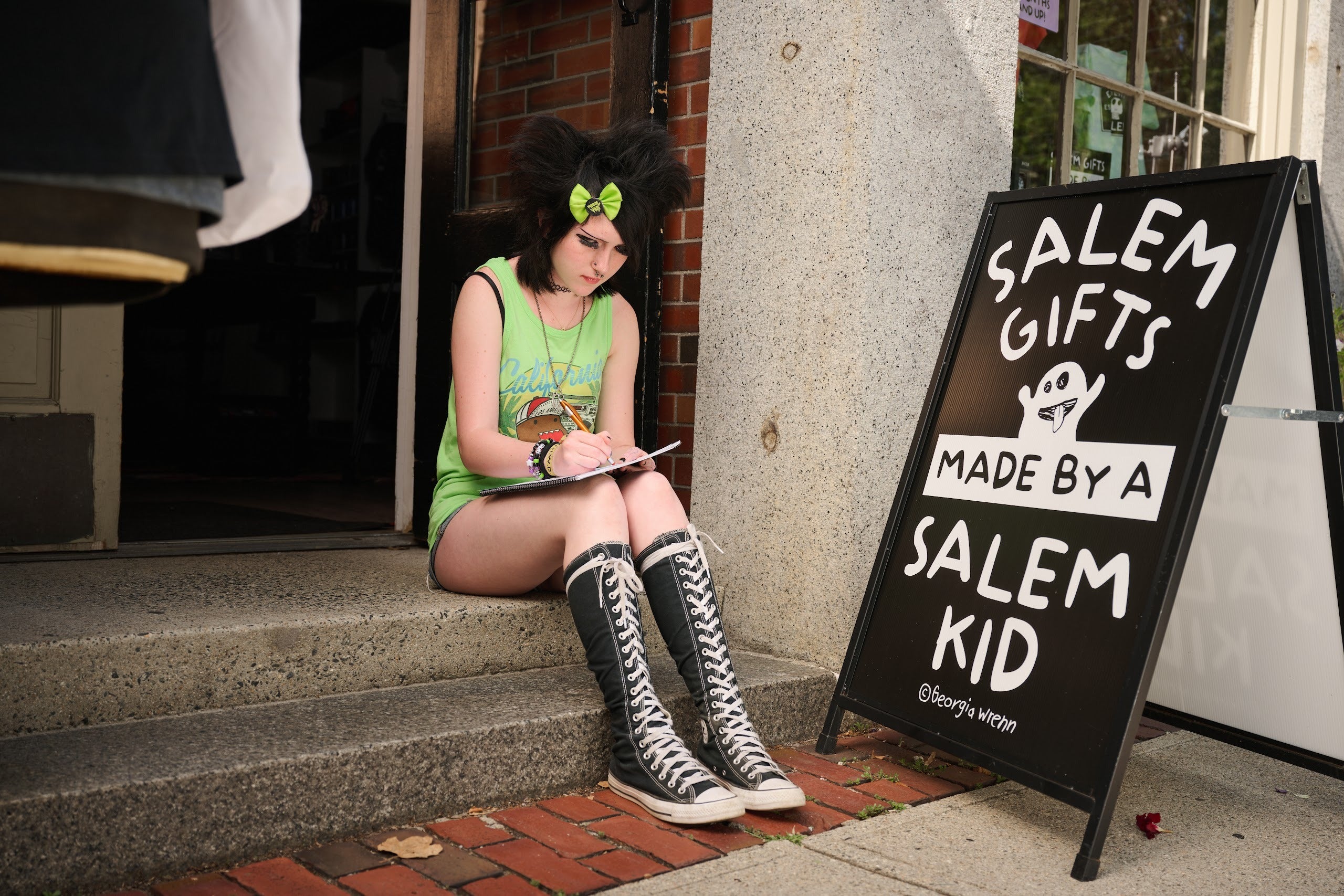 Georgia Wrenn drawing on the step outside her shop in Salem, beside a sandwich board reading Salem Gifts Made By A Salem Kid.